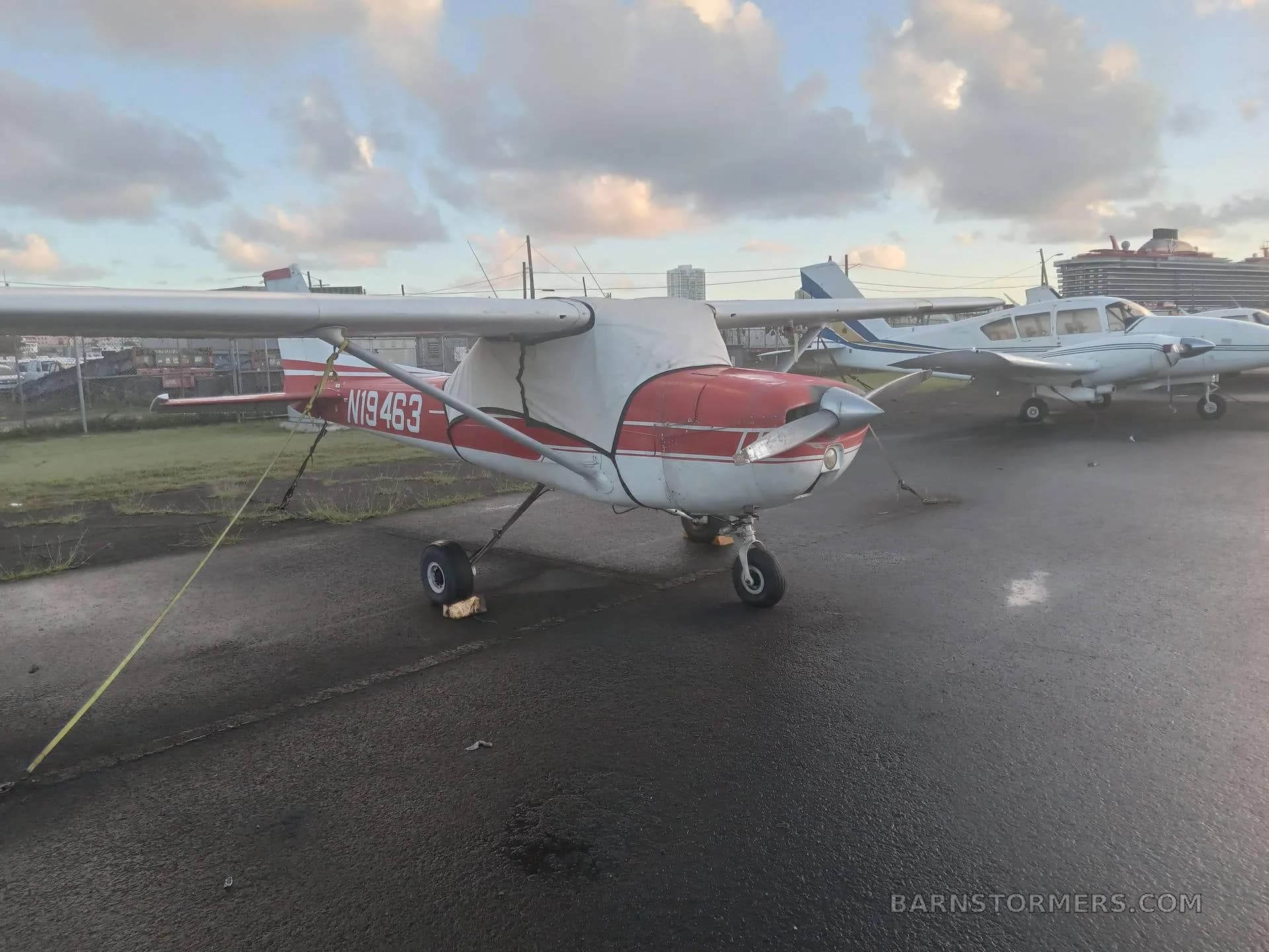 A red and white Cessna 150 (N19463) parked on a wet tarmac with an engine cover, with other aircraft and a cruise ship in the background.