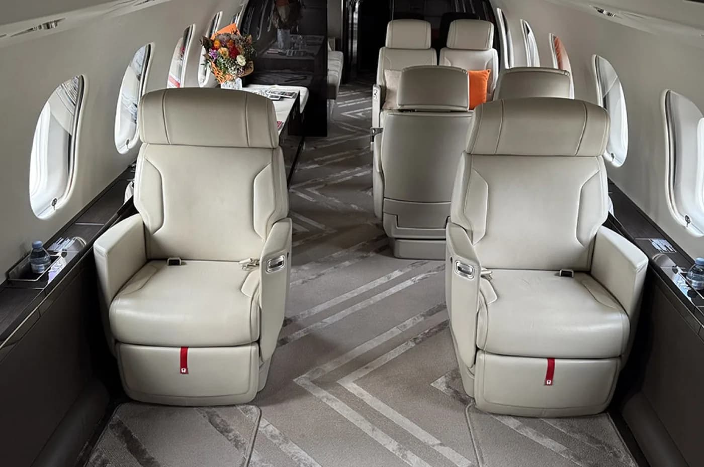 The luxurious cabin interior of a private jet, showing two rows of light-colored leather seats facing each other, with a patterned carpet.