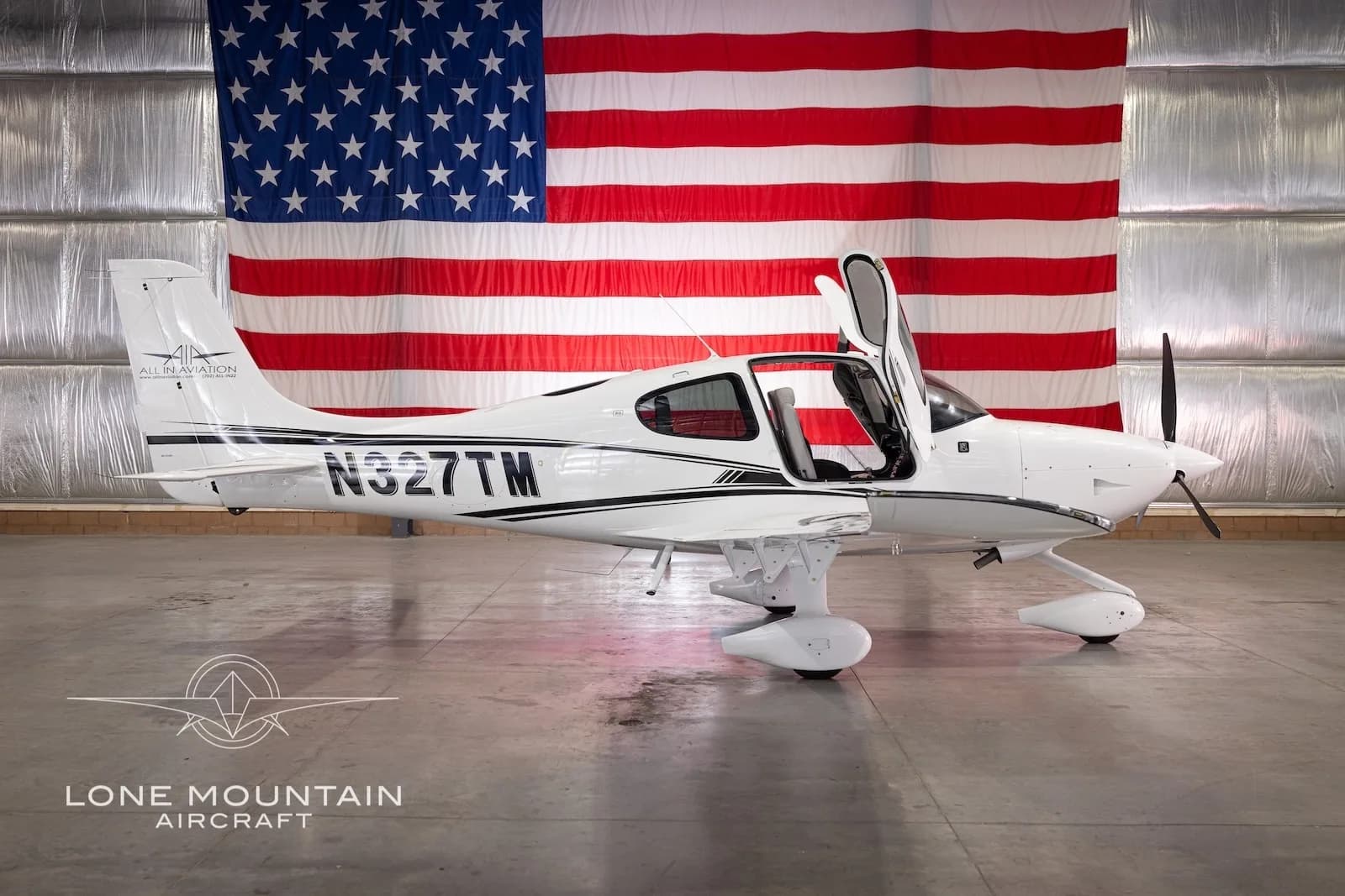 Side view of a white Cirrus SR22 aircraft with black stripes and registration N327TM, parked in a hangar with an American flag backdrop, with its gull-wing doors open.