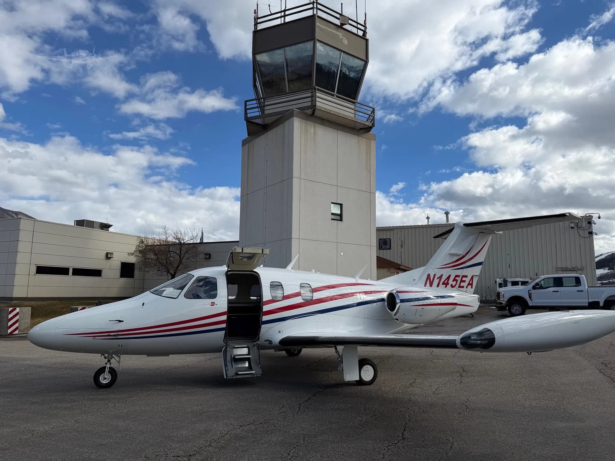 A white Cessna CitationJet with red and blue stripes is parked on an asphalt tarmac next to an air traffic control tower under a partly cloudy sky.