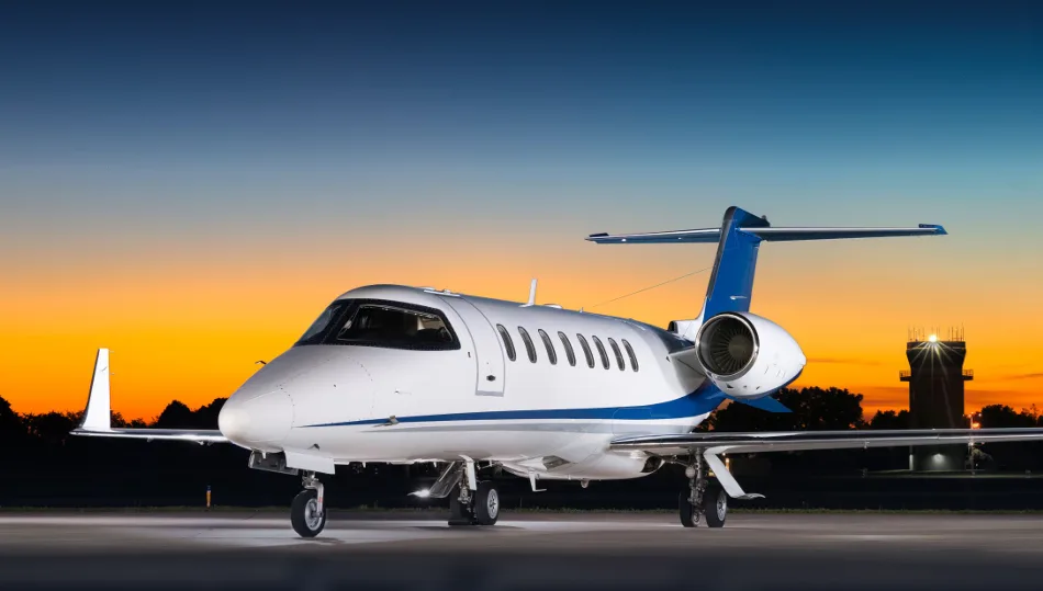 A white and blue business jet is parked on the tarmac at sunset, with an airport tower in the background.