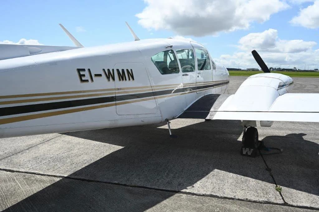 A close-up view of the white fuselage and wing of a twin-engine aircraft with gold and black stripes, registration EI-WMN, parked on a sunny tarmac.