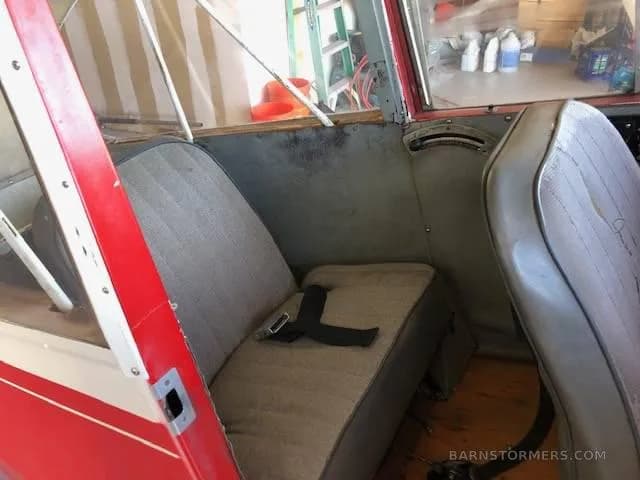 The interior cabin of a light aircraft showing two grey fabric seats with a red and white frame visible.