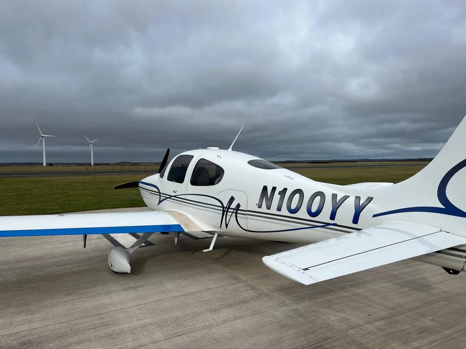 A white Cirrus SR22 aircraft with blue and grey stripes parked on an airfield under a cloudy sky, with wind turbines in the distance.