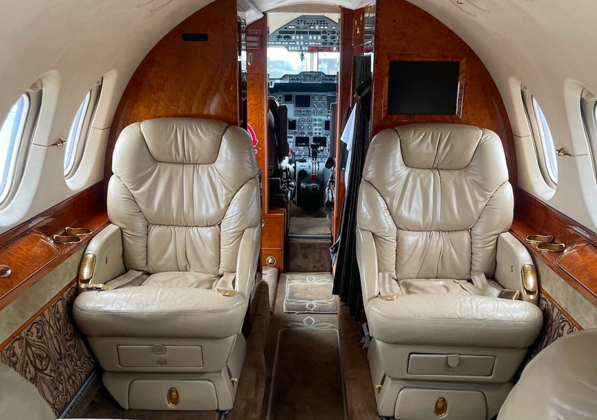 The interior cabin of a business jet with two beige leather seats facing forward, looking towards the cockpit.