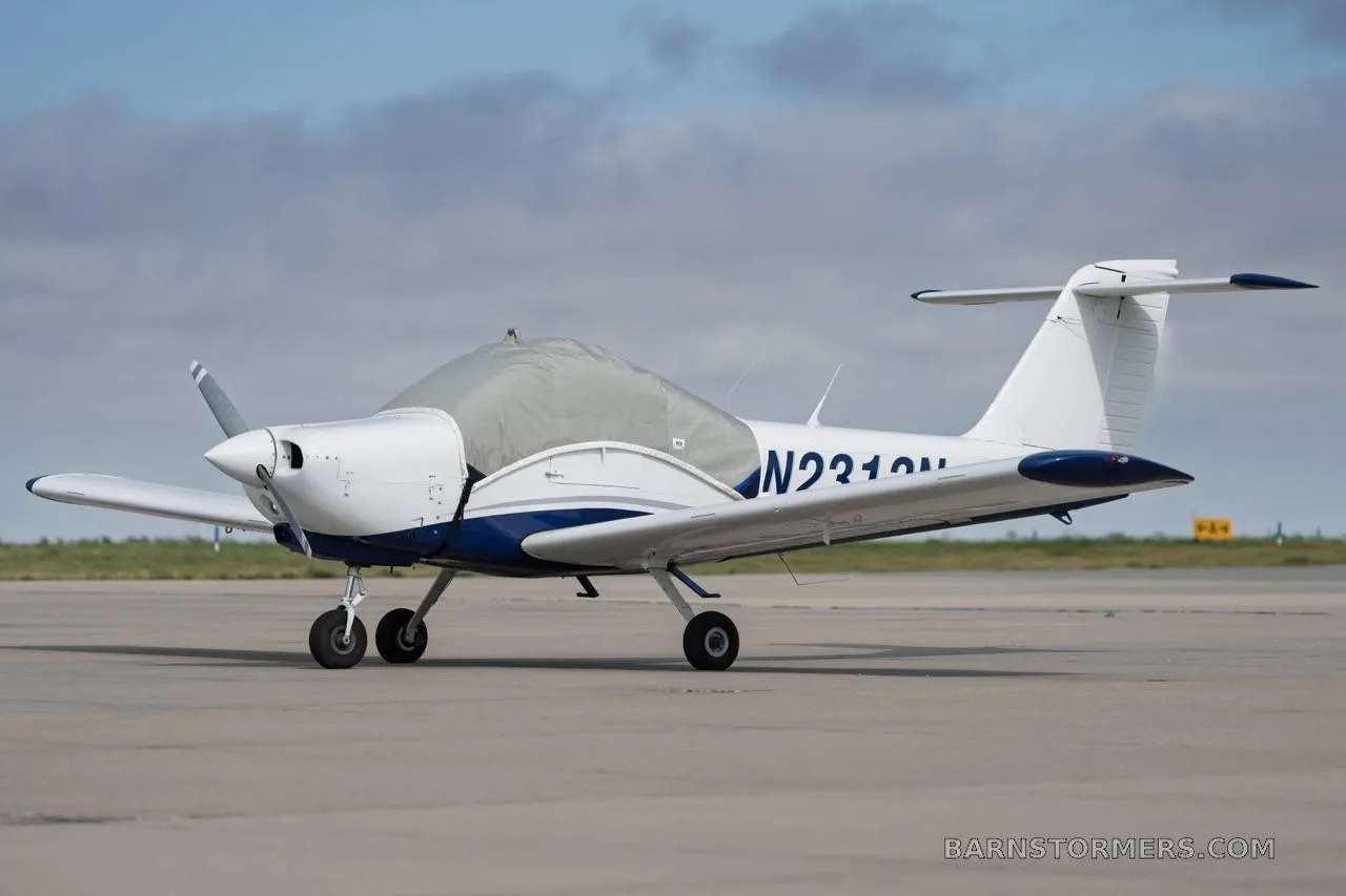 A white and blue single-engine propeller aircraft with a gray cockpit cover is parked on an asphalt runway under a cloudy sky, viewed from the side.