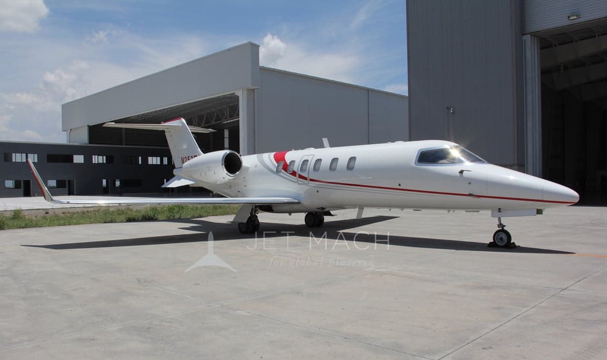 A white Learjet 75 with red and grey stripes parked on a concrete apron outside a hangar.