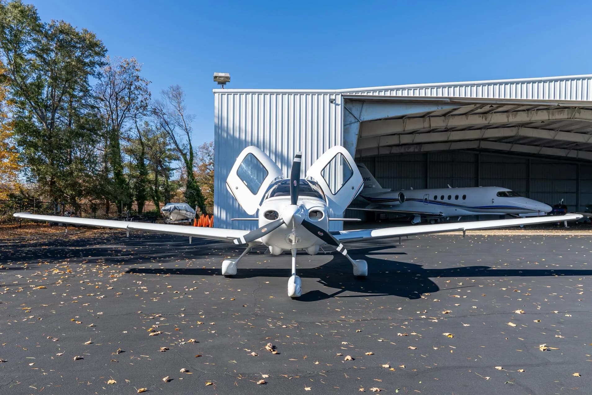 A white Cirrus SR20 aircraft with its doors open is parked in front of a hangar, viewed from the front.