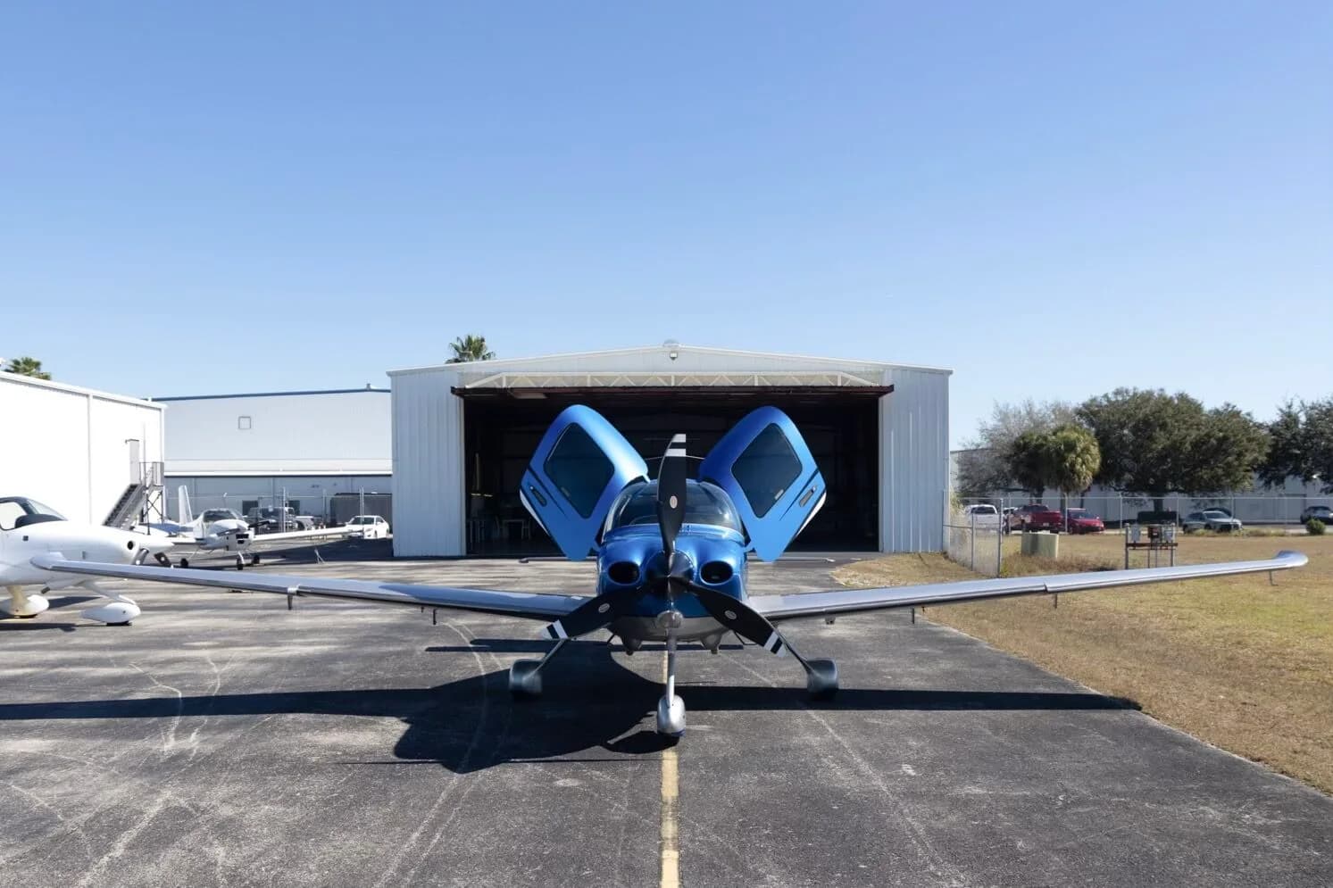 A front view of a blue and silver Cirrus SR22 aircraft (N515SC) with its doors open, parked on an airfield in front of a hangar.