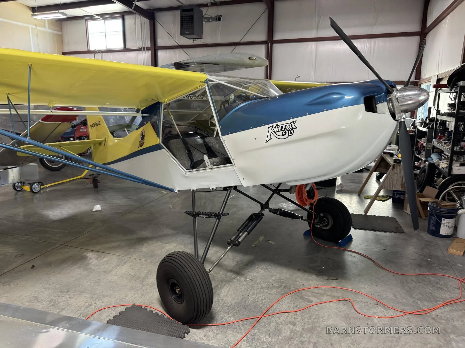 A yellow and blue Kitfox aircraft with its door open, parked in a hangar.