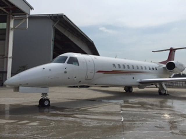 A white business jet with red and gold stripes on the fuselage, parked on wet tarmac next to a hangar.