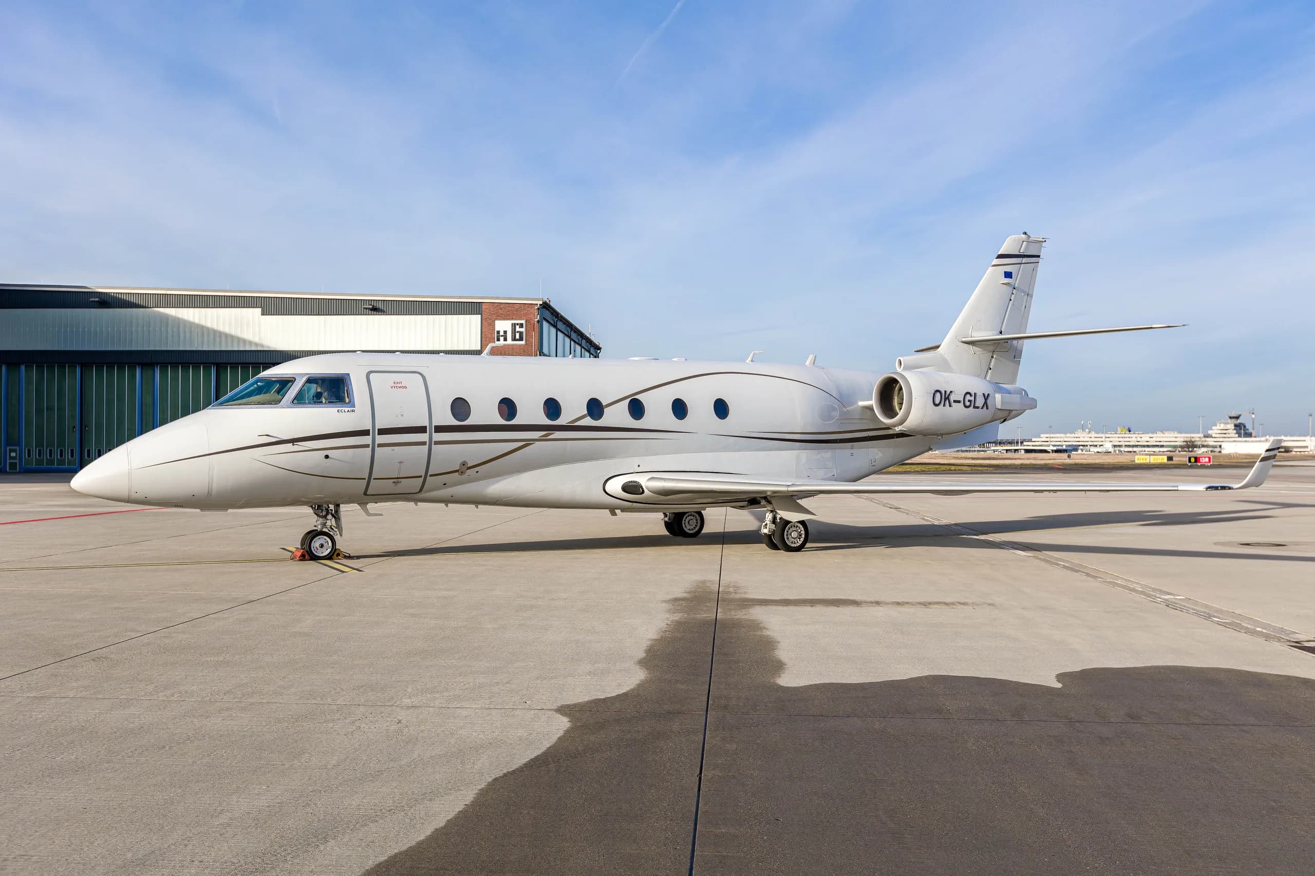 A white Gulfstream G200 business jet with brown accent stripes and registration OK-GLX parked on an airport tarmac.