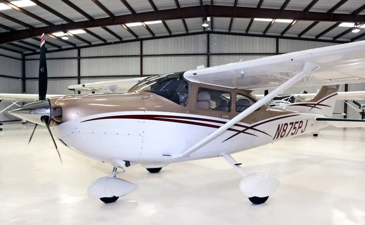 A front-side view of the Cessna Skylane N875PJ, painted brown and white, inside a hangar.