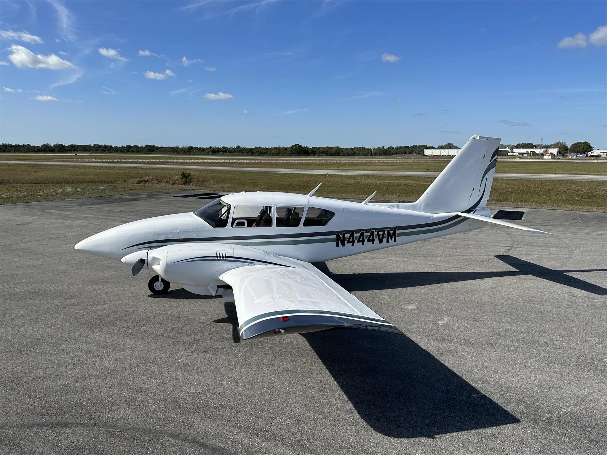 A side view of a white Piper aircraft with green and black stripes, registration N444VM, parked on an airfield.