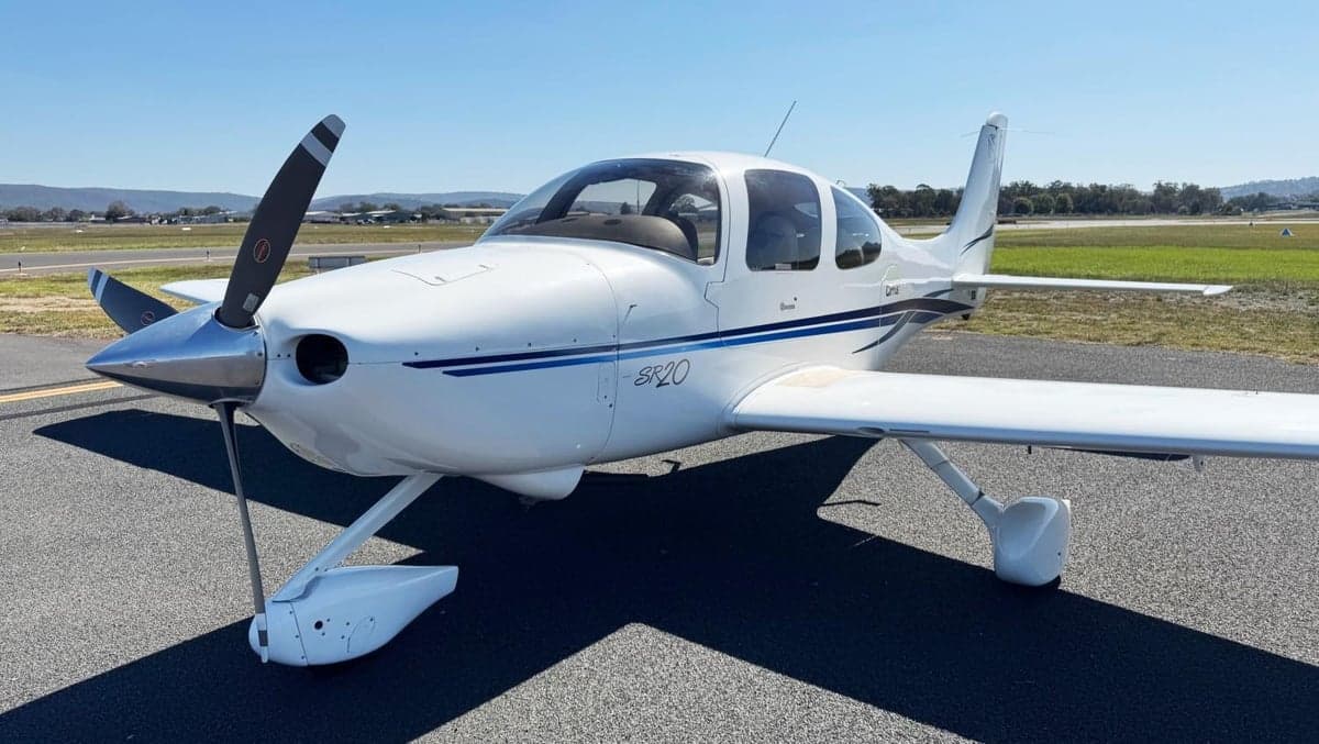 A front-quarter view of a white Cirrus SR20 aircraft with blue stripes, parked on an airfield.