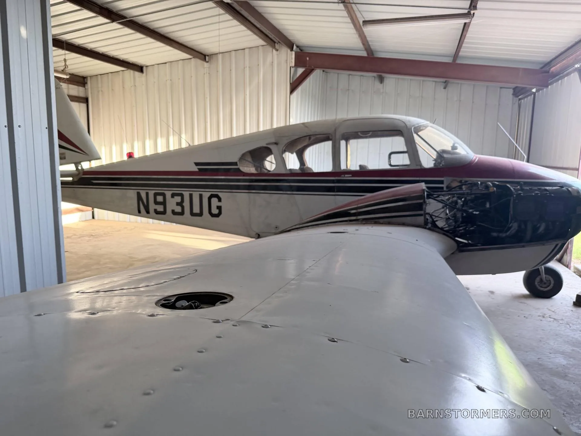 A white and maroon single-engine aircraft with its engine cowling open, parked inside a hangar.