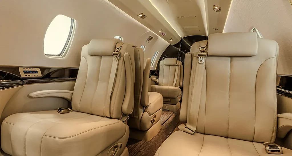 A view inside the private jet cabin, showing multiple beige leather seats and a dark wood-paneled side.