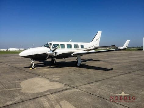 A white and dark-colored twin-engine propeller aircraft with a gold stripe is parked on an airport tarmac under a clear blue sky.