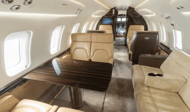 The luxurious interior of a business jet cabin with tan leather seats, a dark wood table, and ambient lighting.