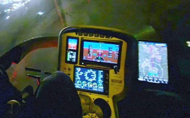 A modern helicopter cockpit with multiple digital displays showing flight information and a navigation map, seen at night.