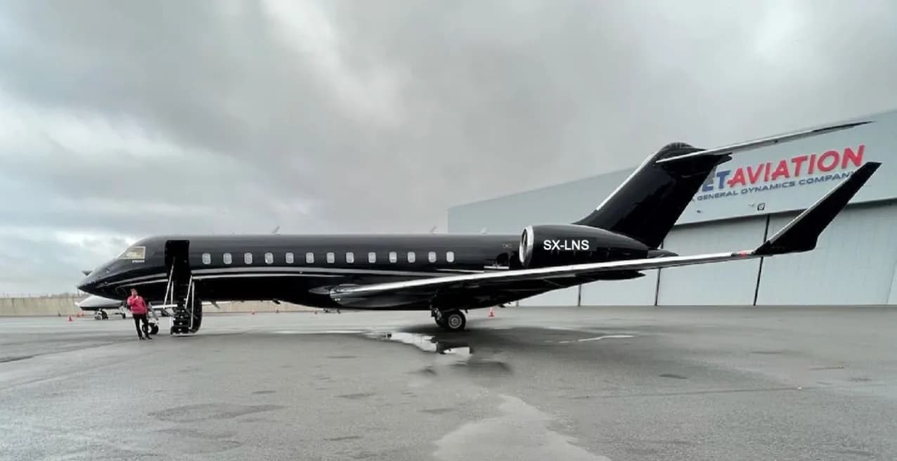 A black Bombardier Global Express XRS aircraft with registration SX-LNS parked on a wet tarmac in front of a hangar, with a person near the open door.