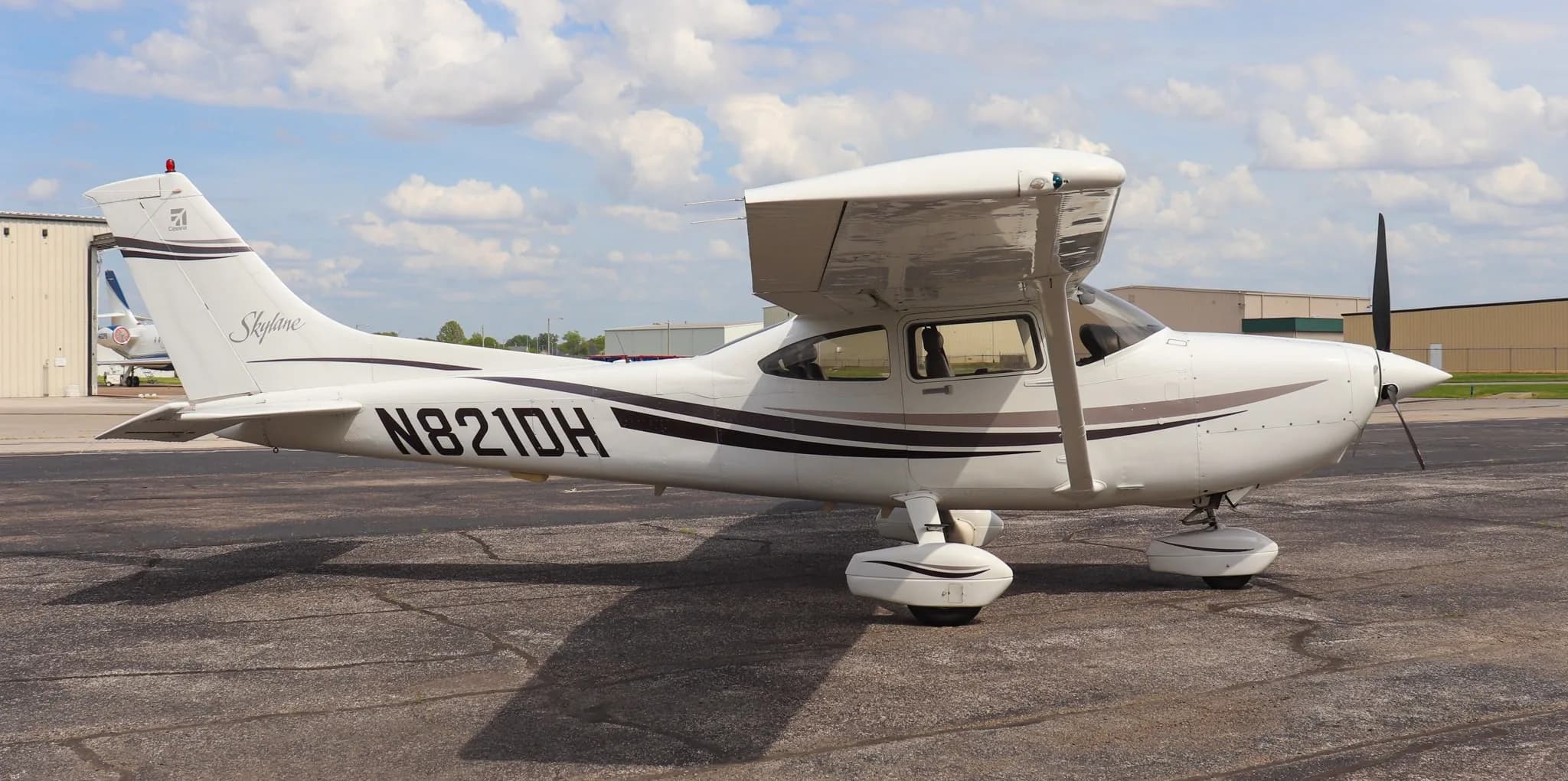 A side view of the white Cessna Skylane N821DH with dark grey and brown stripes, parked on an asphalt tarmac.
