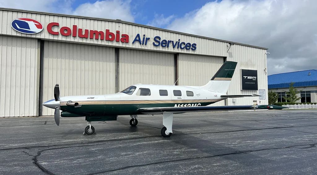 A white and dark green single-engine turboprop aircraft, N110MC, parked in front of a Columbia Air Services hangar.