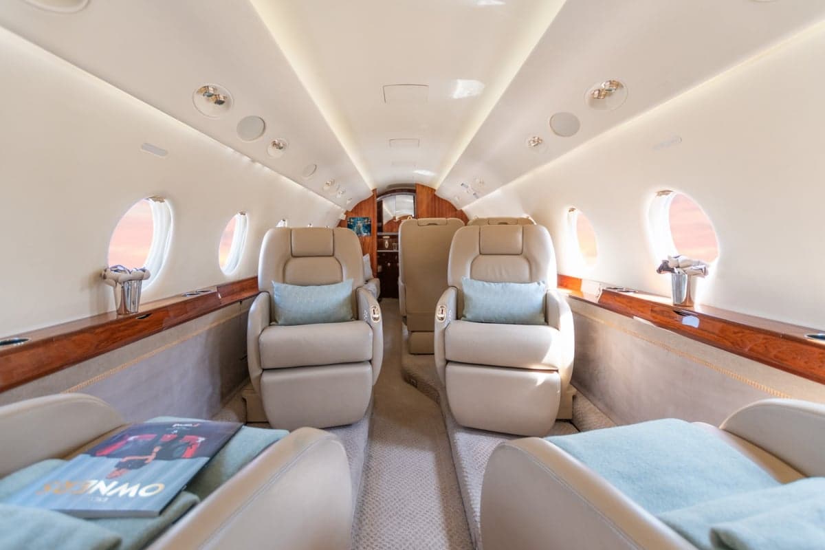 A luxurious aircraft cabin interior with two rows of beige leather seats facing each other, a wooden side table, and large windows.