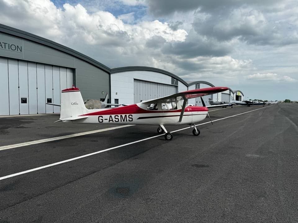 A white and red Cessna 150 G-ASMS parked on the tarmac, viewed from the rear-left, with hangars in the distance.