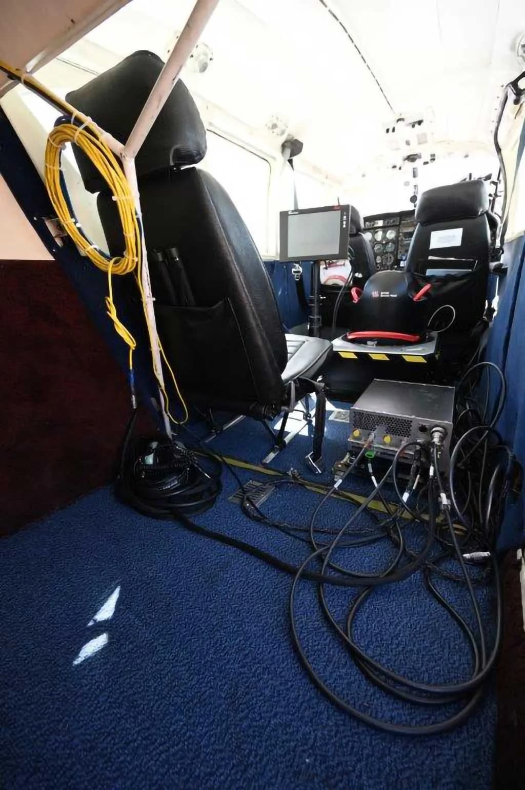 The interior of an aircraft cabin with blue carpet, black seats, and numerous cables and equipment, looking towards the cockpit.