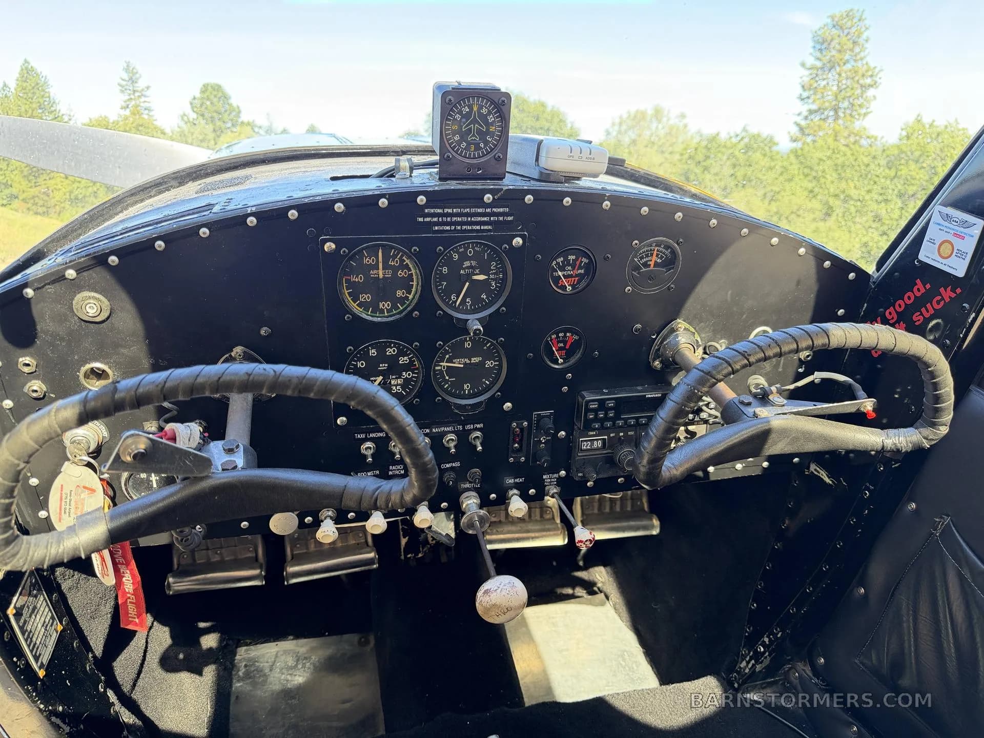 The black instrument panel of a vintage aircraft features dual yokes, various gauges, and a radio, with a propeller visible through the windshield.
