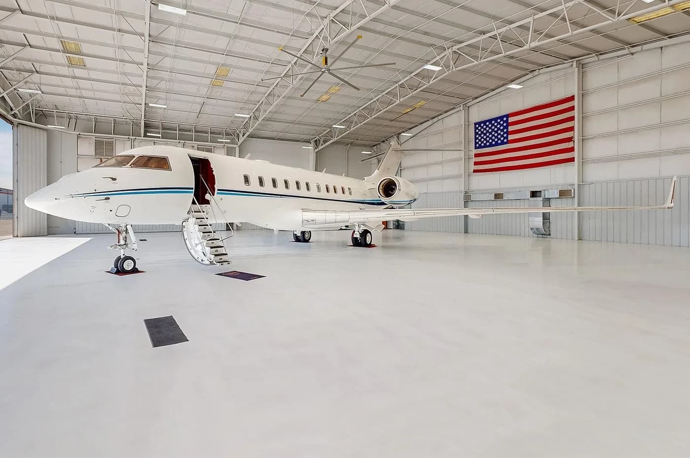 A white business jet with blue stripes parked in a brightly lit hangar with an American flag on the wall, seen from a slightly different angle.