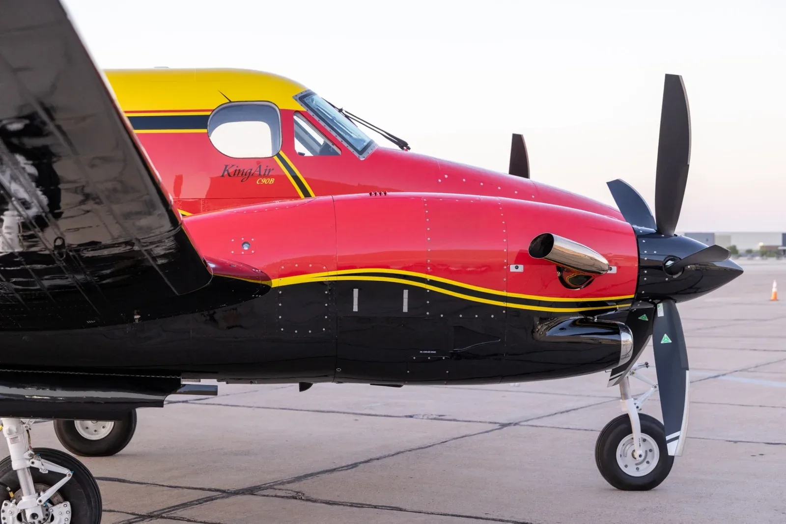A close-up side view of the front fuselage and engine of a red, yellow, and black King Air C90B.