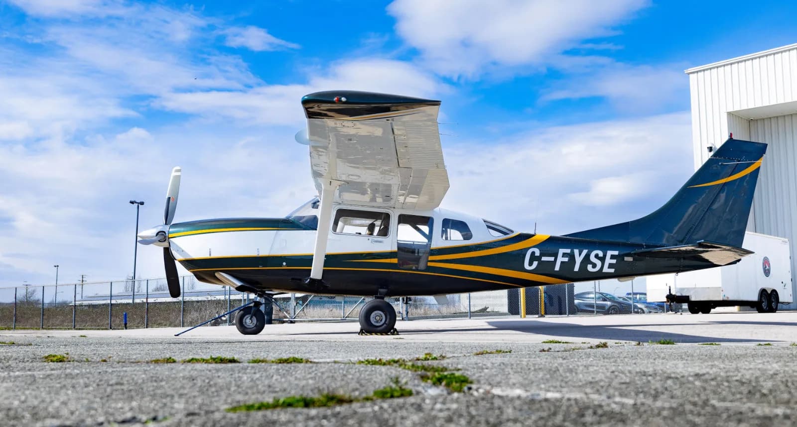 A dark green and white high-wing single-engine aircraft with yellow stripes (C-FYSE) is parked on an asphalt tarmac next to a white hangar under a blue sky.