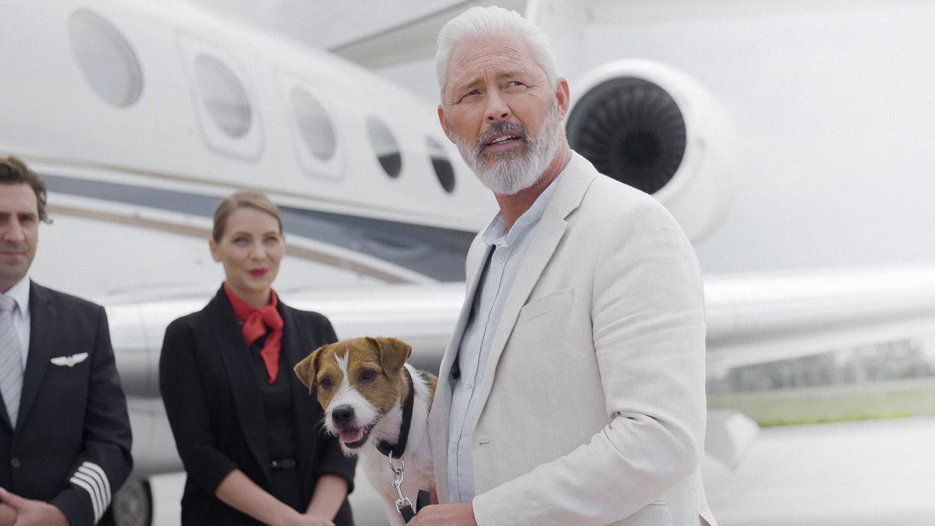 A man holding a small dog, standing with a pilot and flight attendant in front of a white business jet.