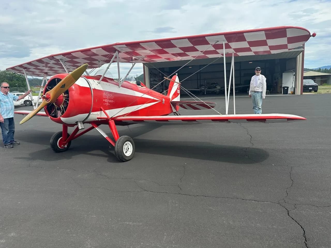 A red and white biplane with a checkered upper wing, parked on an asphalt apron with two people standing near it.