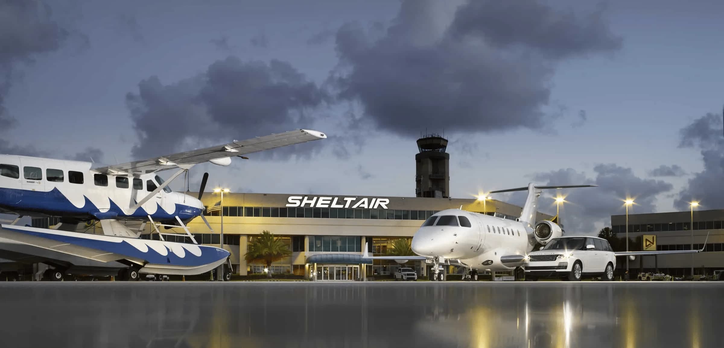 A seaplane and a business jet are parked on a wet tarmac in front of the Sheltair FBO building at dusk.