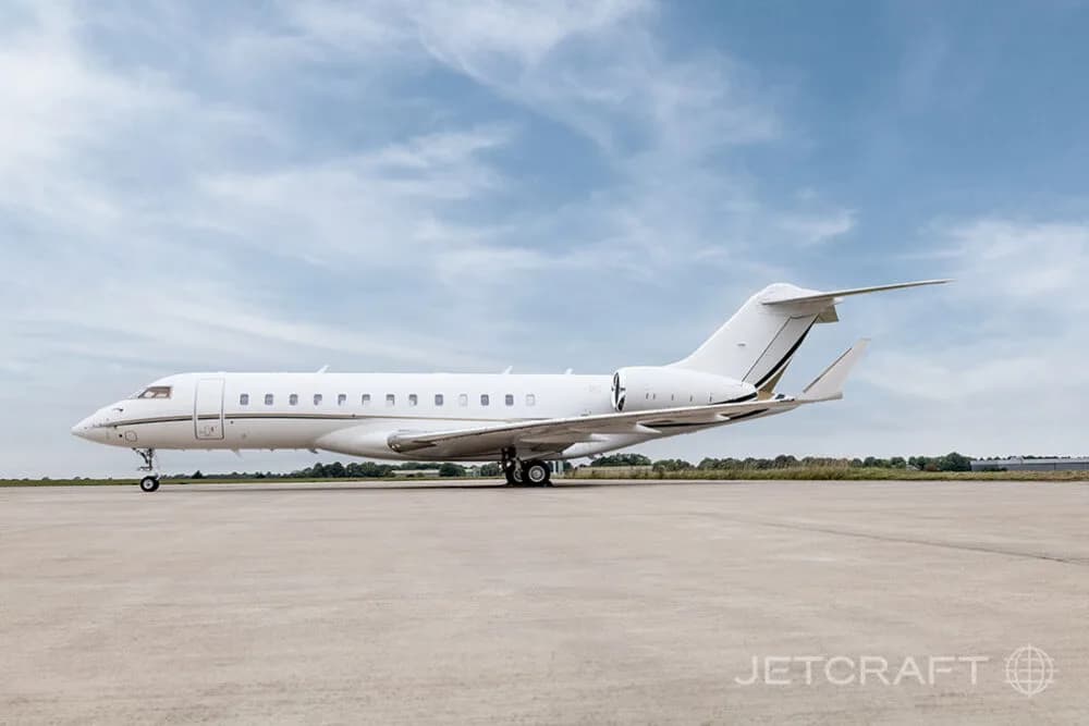 A white Bombardier Global business jet with gold and black stripes parked on an airfield, viewed from the side.