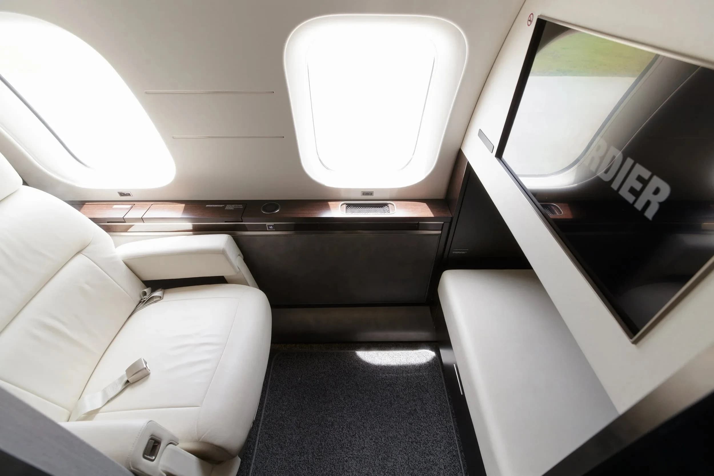 A plush white leather seat and a large screen displaying "BOMBARDIER" are visible in the aircraft cabin.