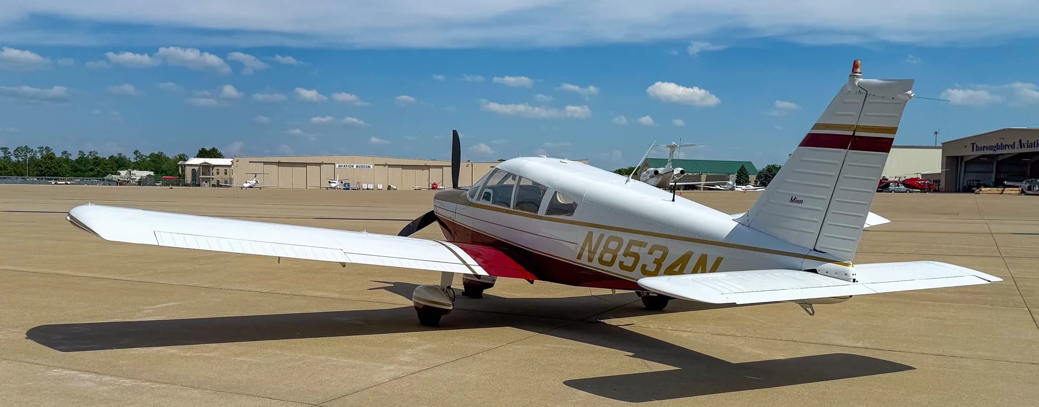 A three-quarter rear view of the white, red, and gold Piper Cherokee aircraft (N8534N) on an airfield.