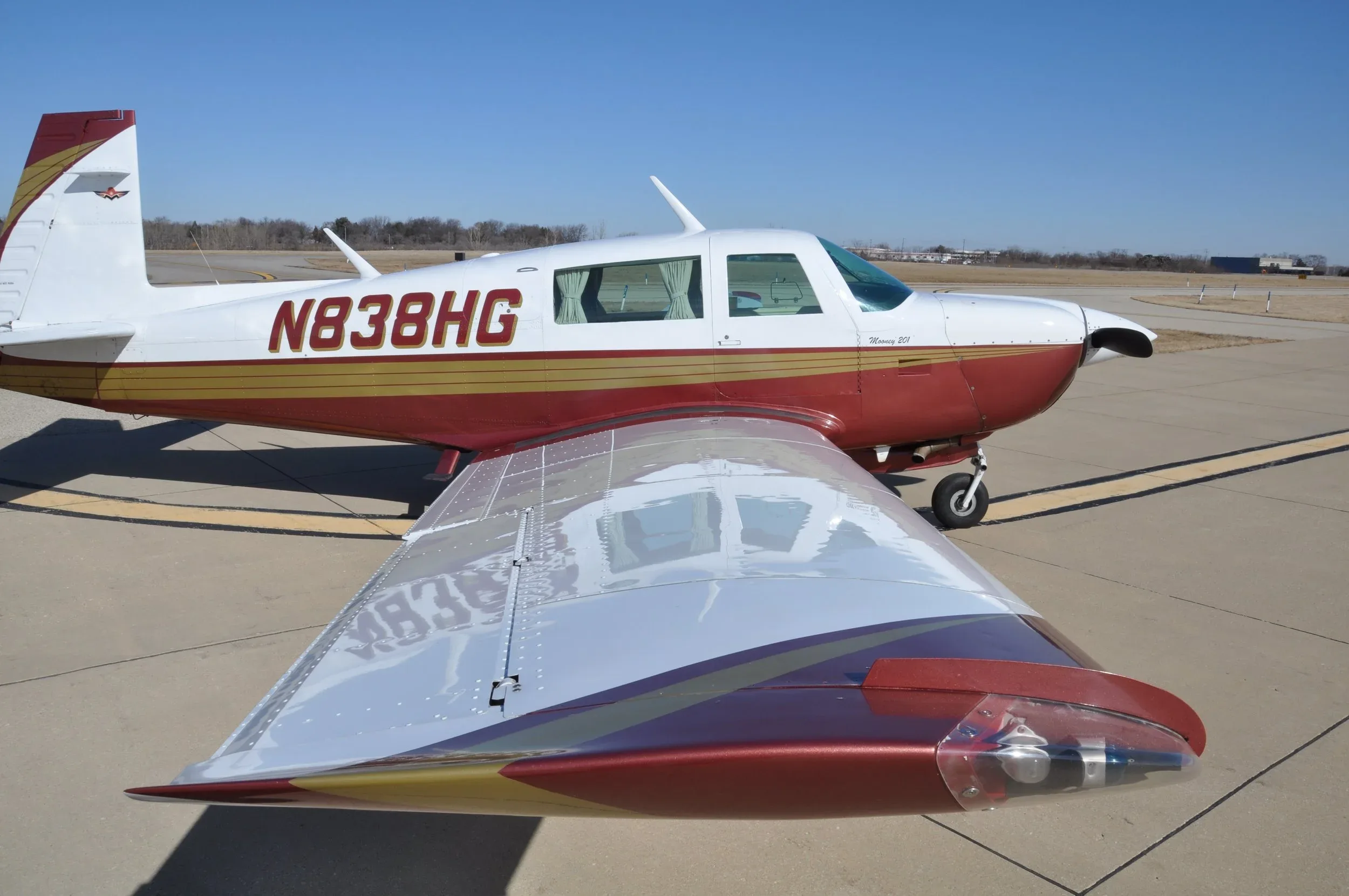 A close-up side view of a red and white Mooney 201 aircraft with registration N838HG, showing its wing and fuselage on an airfield.