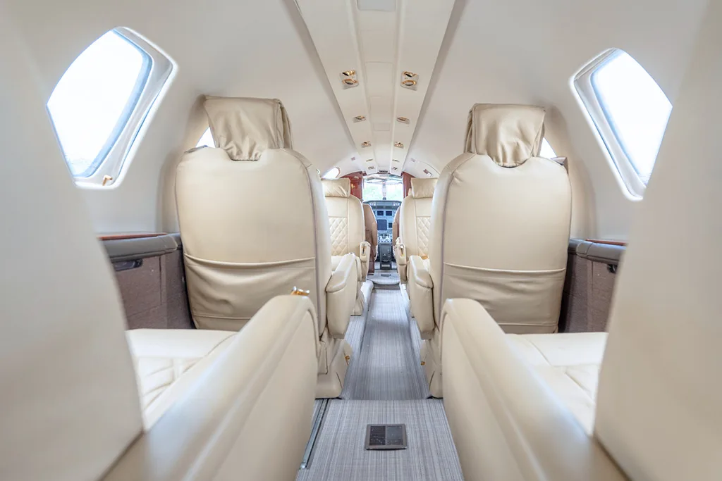 A view down the aisle of a private jet cabin with light beige seats, looking towards the cockpit.