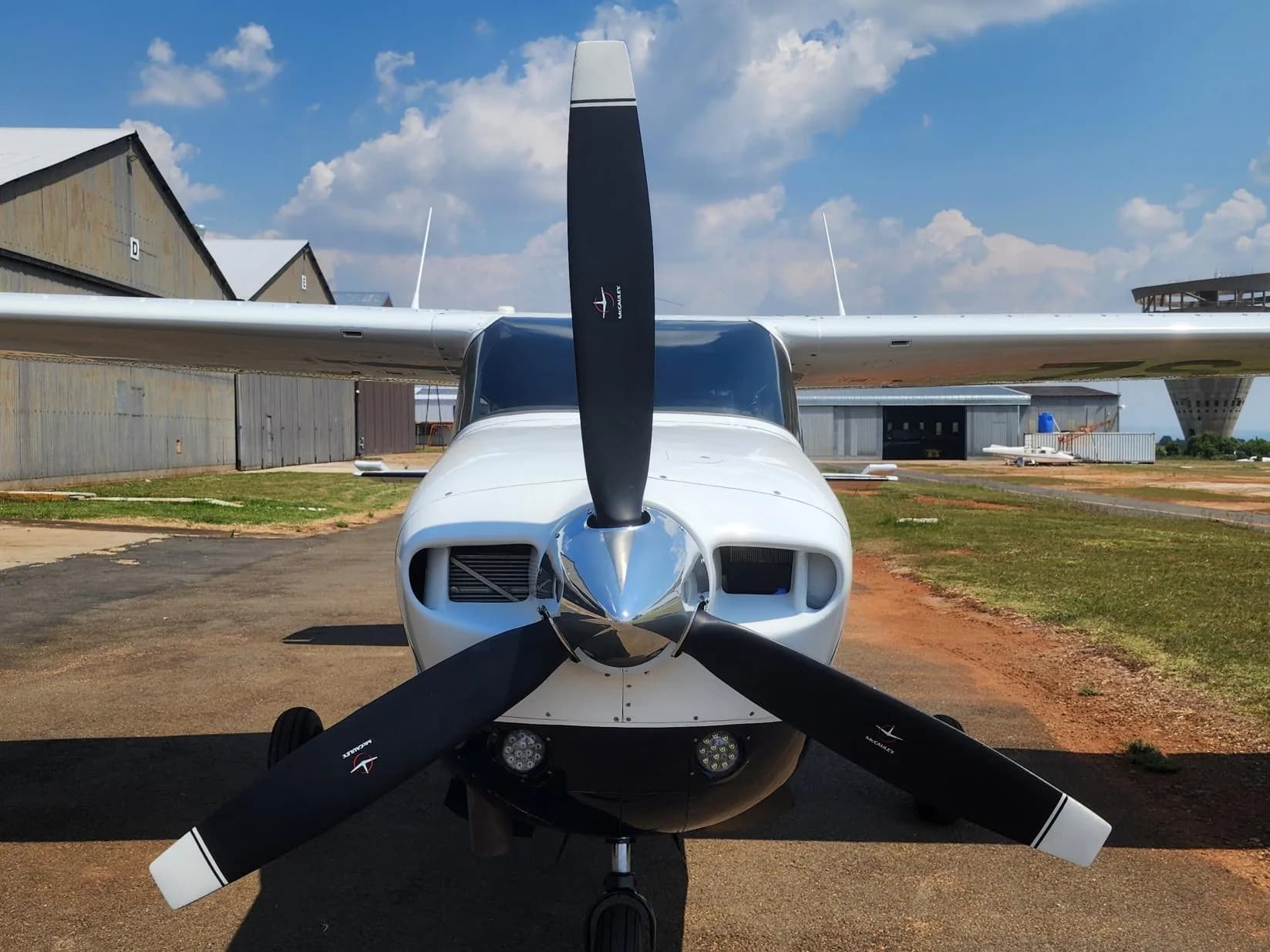 A white single-engine propeller aircraft with a black propeller is parked on an airfield, viewed from the front.