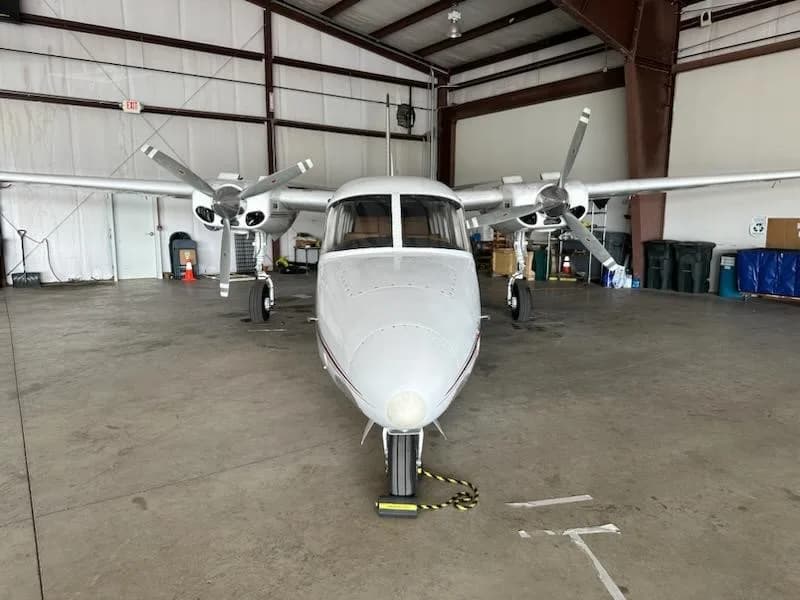 A head-on view of a white Aero Commander 680FL twin-engine aircraft parked in a hangar.