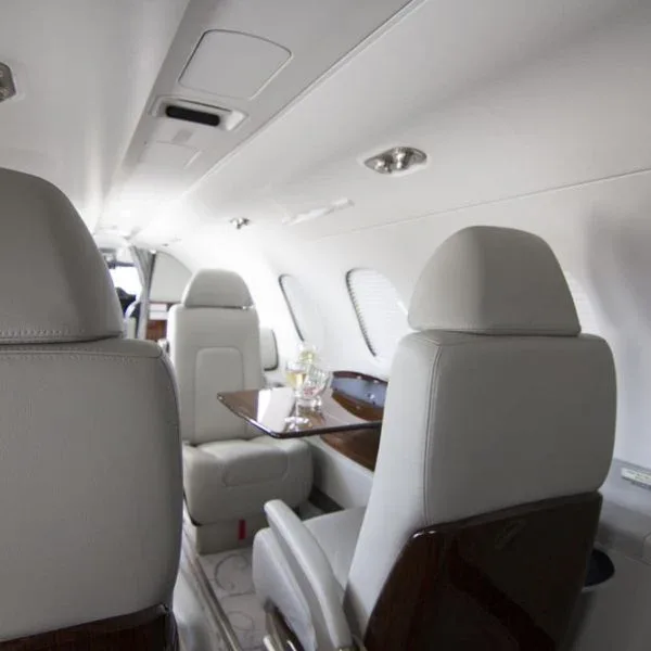 A private jet cabin interior showing light grey seats and a small table with drinks.
