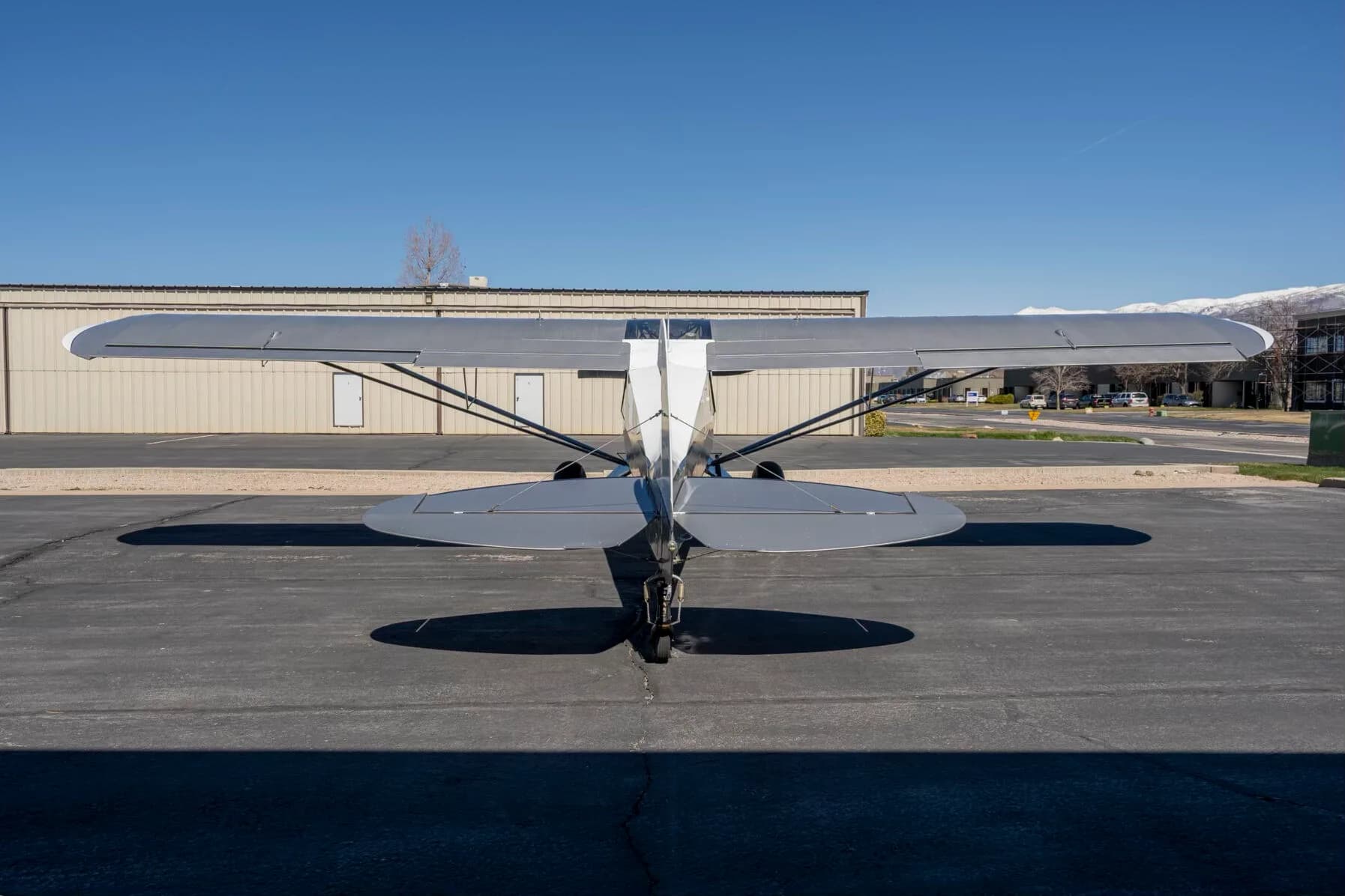 A rear view of the white and grey Carbon Cub FX-3 aircraft N25FX parked on an asphalt ramp in front of a hangar.