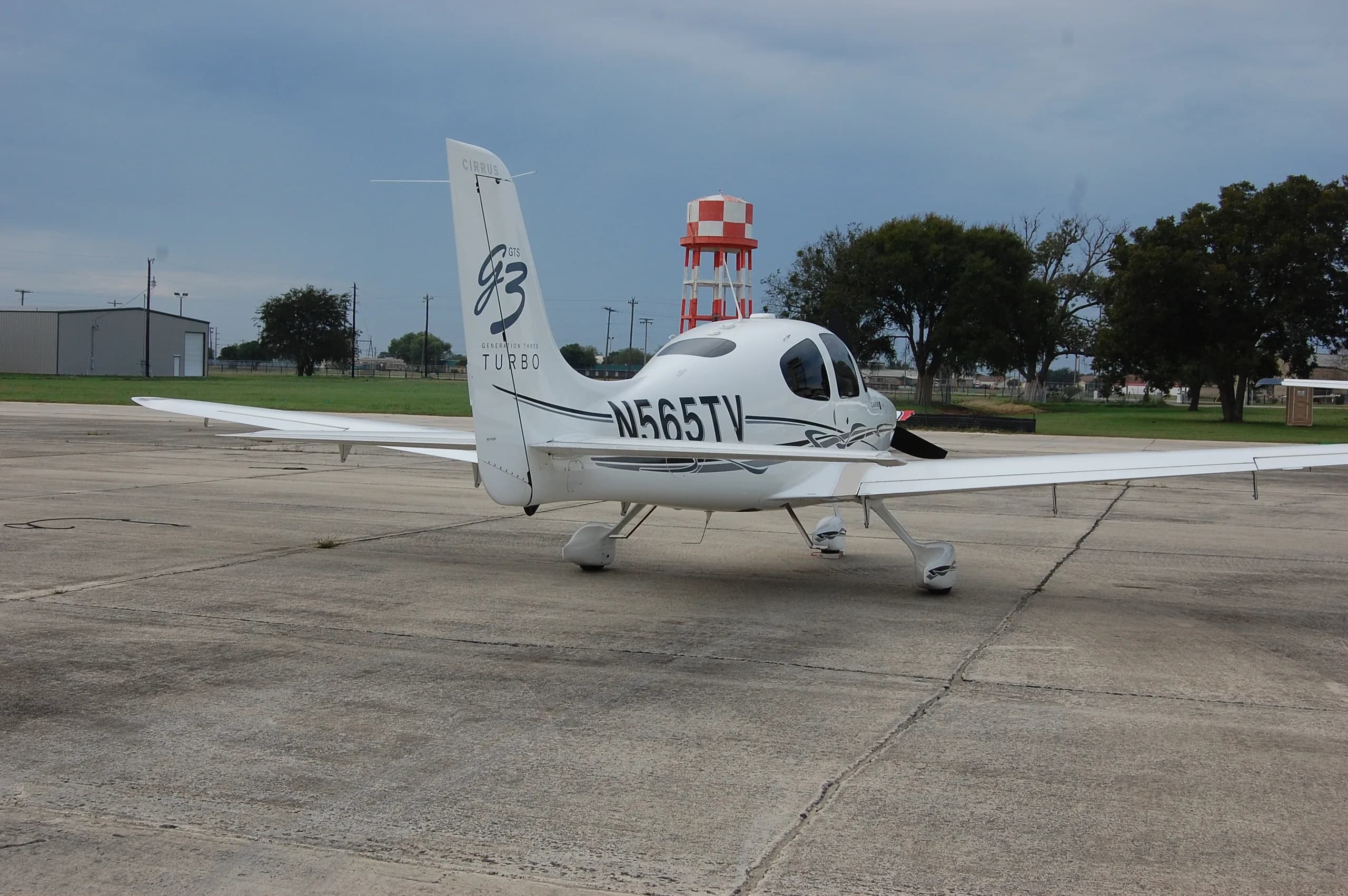 A rear-quarter view of the white Cirrus SR22T aircraft, N565TV, parked on a tarmac with a red and white water tower in the background.
