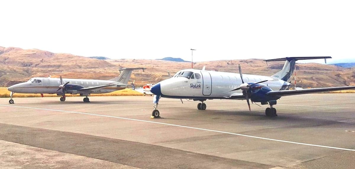Two turboprop aircraft, one white and blue SkyLink and one grey, parked on an airport tarmac with hills in the background.