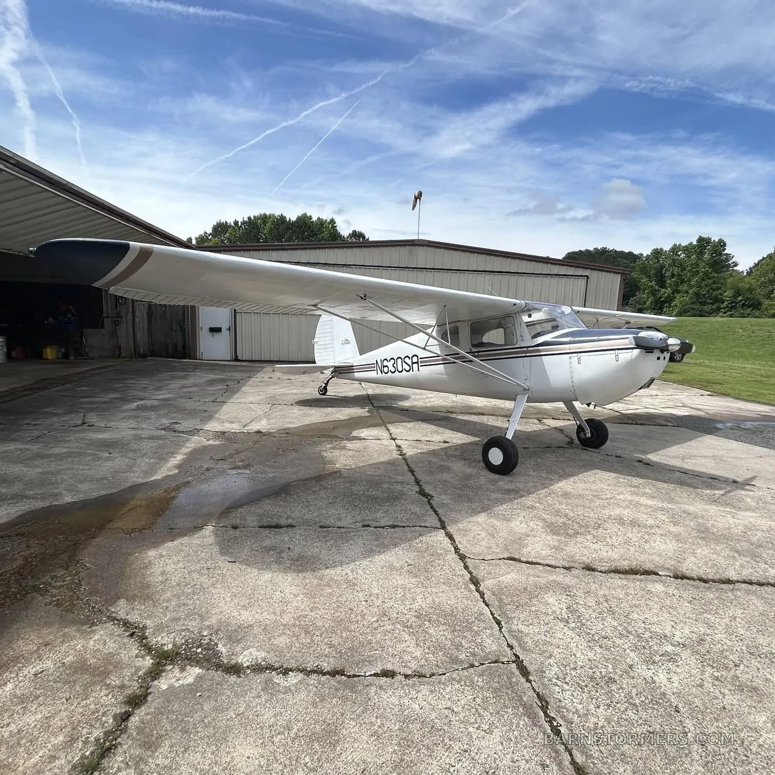 A white Cessna 170B with dark blue and gold stripes is parked on a concrete pad next to a hangar under a blue sky with contrails.