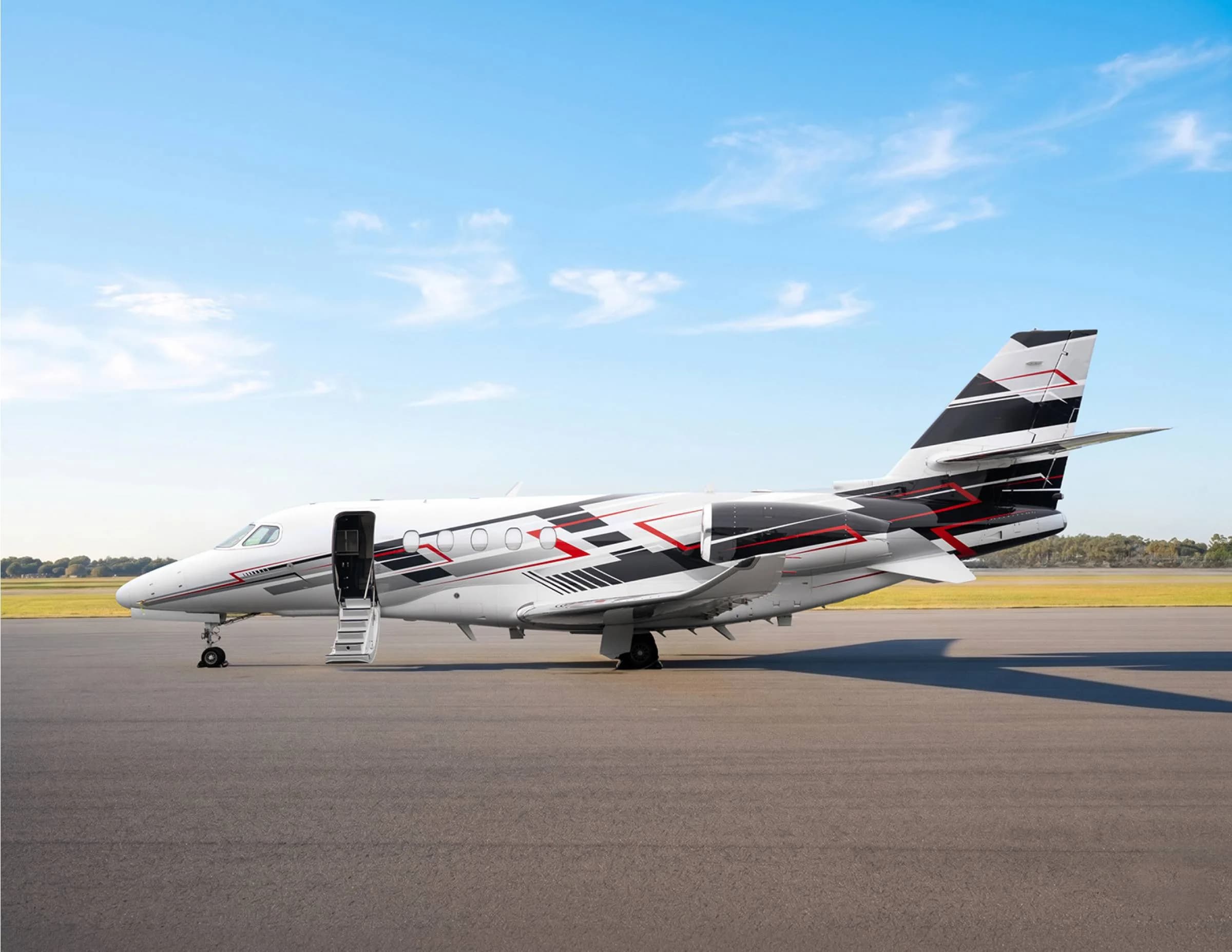 A side view of a white private jet with black and red geometric livery parked on an asphalt runway under a blue sky.